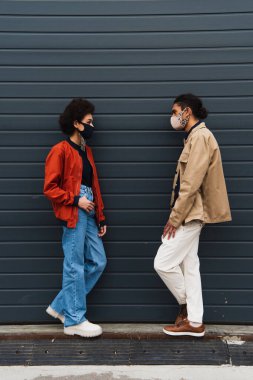 Tam boy Afro-Amerikan kadın ve koruyucu maskeli şık bir adam dışarıda birbirlerine bakıyorlar. 