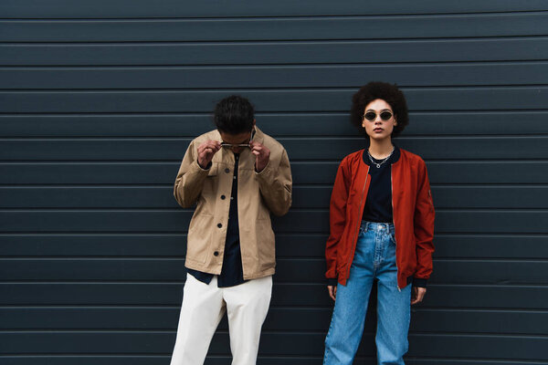 young african american man adjusting sunglasses and posing with stylish woman in jeans outside 