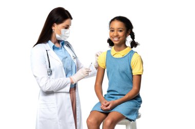 Tıbbi maskeli doktor elinde şırıngayla beyaz tenli, neşeli Afrikalı Amerikalı bir çocuğun yanında. 