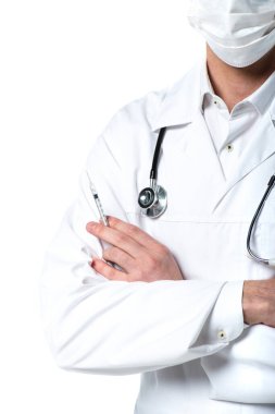 Tıbbi maskeli doktorun elinde şırıngayla çapraz kollarıyla beyazın üzerinde dikilirken görüntüsü. 