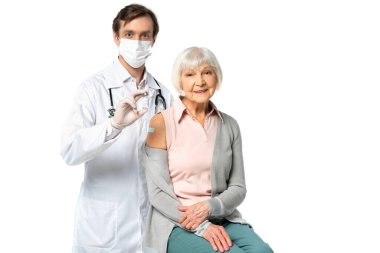 Tıbbi maskeli doktor aşıyı elinde tutuyor. Yanında gülümseyen bir hasta var. 
