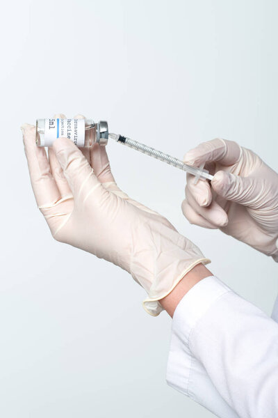 Cropped view of doctor in latex gloves picking up coronavirus vaccine in syringe isolated on grey