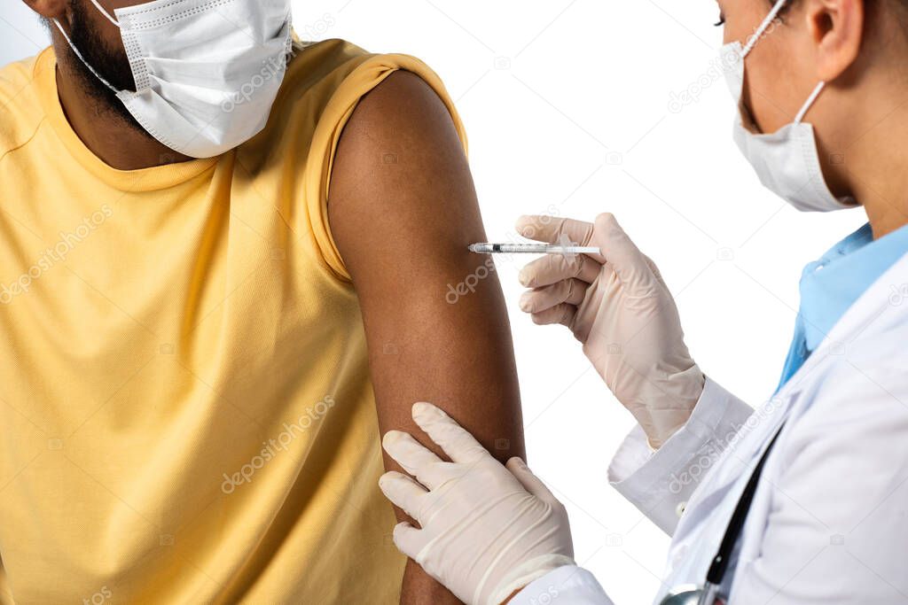 African american patient in protective mask near doctor doing vaccination on blurred foreground isolated on white 