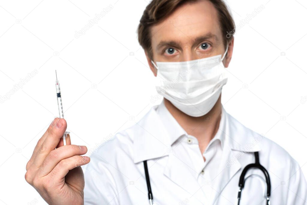 Syringe in hand of blurred doctor in medical mask isolated on white 