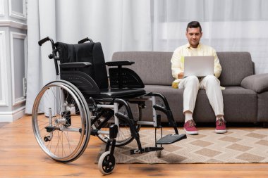 disabled man working on sofa with laptop near wheelchair on foreground clipart