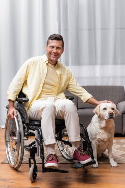 Labrador köpeğini okşarken kameraya gülümseyen tekerlekli sandalyedeki engelli adam.