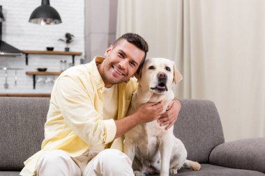 Labrador köpeğini kucaklarken kameraya gülümseyen mutlu adam.