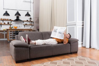 Genç adam gazete okurken modern dairede kanepede yatıyor.