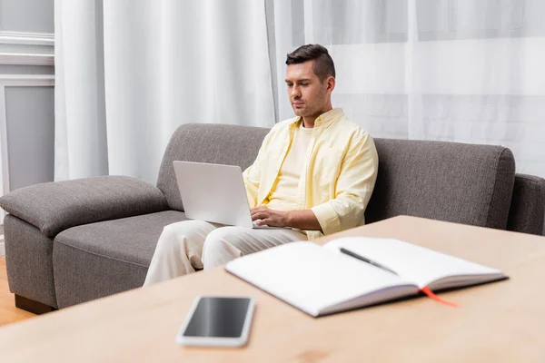 young man working on laptop near notebook and mobile phone on blurred foreground