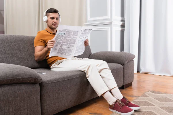 Fiatal Férfi Olvas Újságot Miközben Zenét Hallgat Fejhallgató Kanapén Otthon — Stock Fotó