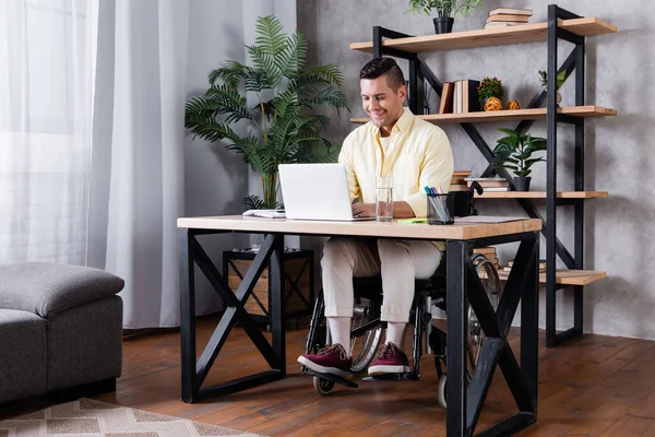 positive handicapped man in wheelchair working near laptop at home