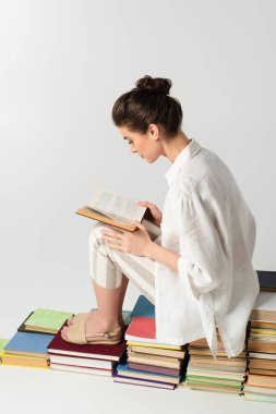 Genç esmer kadın, beyazın üzerinde izole bir şekilde kitap yığını üzerinde otururken kitap okuyor. 