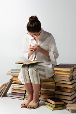 Uzun boylu, gözlüklü esmer bir kadın kitap yığınının üzerinde oturuyor ve beyaz bardakları okuyor ve tutuyor. 