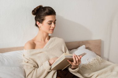 brunette young woman reading book while resting on bed at home clipart