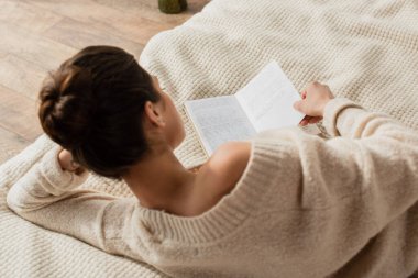 Evdeki yatakta dinlenirken süveter giymiş, kitap okuyan esmer bir kadının arka görüntüsü.