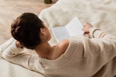 back view of young brunette woman in sweater reading book while resting on bed at home clipart