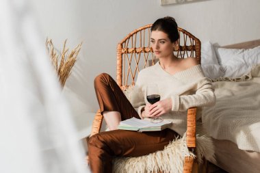 Hasır sandalyede oturan genç ve güzel bir kadın. Elinde kitap ve bir bardak şarapla sallanıyor.