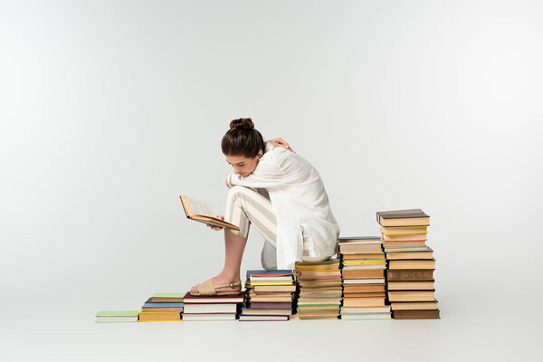 full length of young woman scratching back while sitting on a pile of books on white 