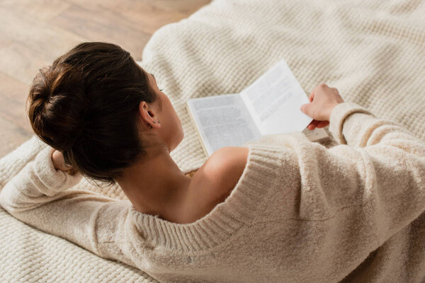 back view of young brunette woman reading book while resting on bed at home