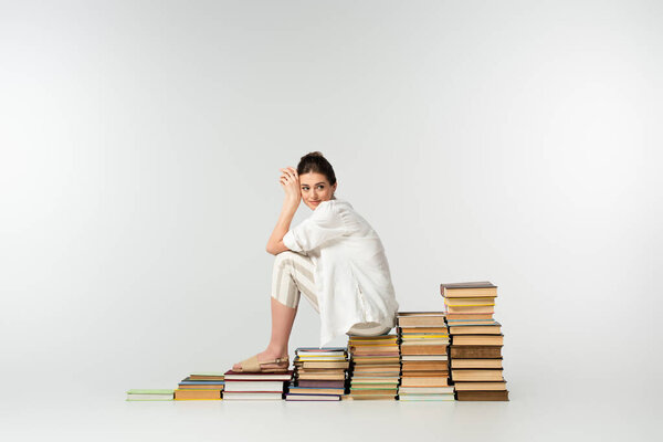 full length of smiling woman in sandals sitting on pile of books on white 