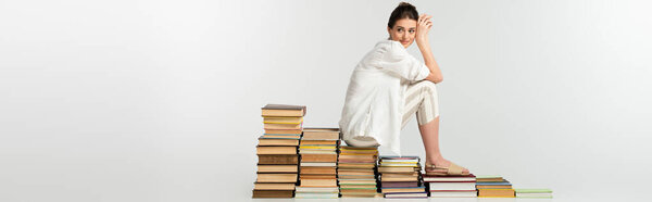 full length of young woman in sandals sitting on pile of books on white, banner
