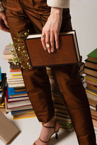 cropped view of young woman holding book in hardcover with flower on grey