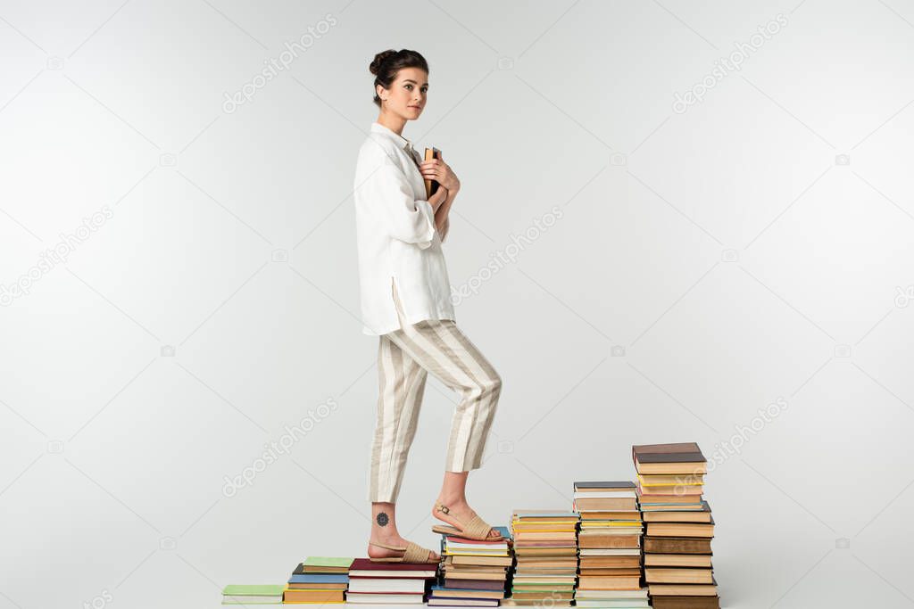 full length of young tattooed woman standing on pile of books isolated on white 