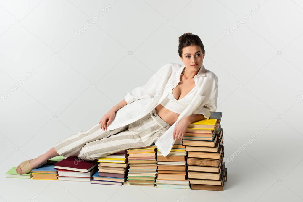 full length of young woman in sandals lying on pile of books on white 