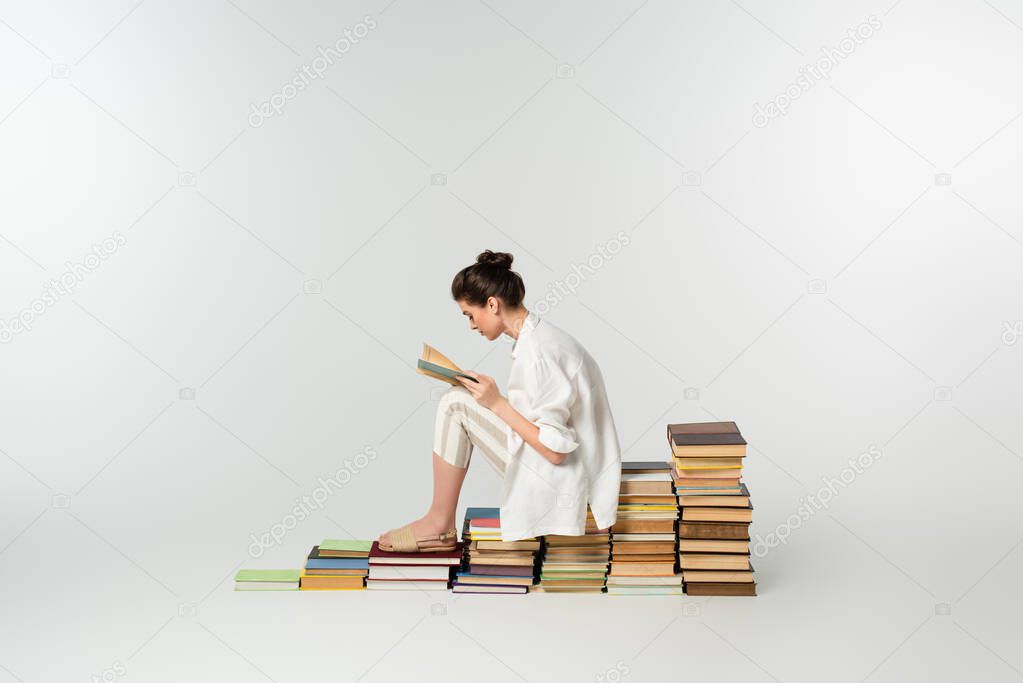 side view of young woman in sandals reading while sitting on pile of books on white 