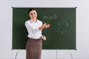 Matematik dersinde gülümseyen öğretmen, tahtaya elinle işaret ediyor. 