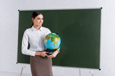 Cheerful teacher holding globe near chalkboard  clipart
