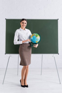 Genç öğretmen elinde küreyle okulda karatahtanın yanındaki kameraya gülümsüyor. 