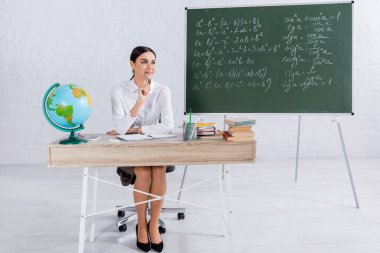 Gülümseyen öğretmen elinde kalemle defter, akıllı telefon ve küreyi tutuyor. 