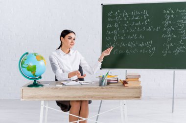 Gülümseyen öğretmen dünyanın yakınındaki tahtaya ve sınıftaki kitaplara işaret ediyor. 