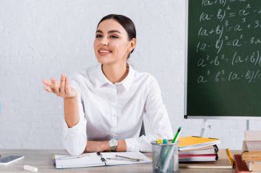 Gülümseyen öğretmen sınıfta kitapların, akıllı telefonun ve tebeşirin yanında oturuyor. 