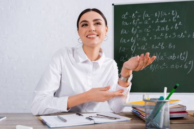 Masanın yanındaki defterleri gösteren olumlu öğretmen. 