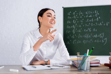 Teacher pointing with finger near notebooks and blurred chalk  clipart