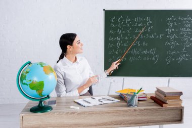Gülümseyen öğretmen okulda kitapların ve dünyanın yakınındaki denklemlerle tahtayı işaret ediyor. 
