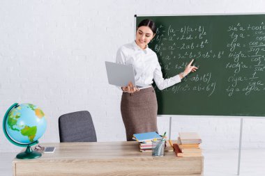 Gülümseyen öğretmen sınıfta bilgisayar başında matematik denklemine işaret ediyor. 