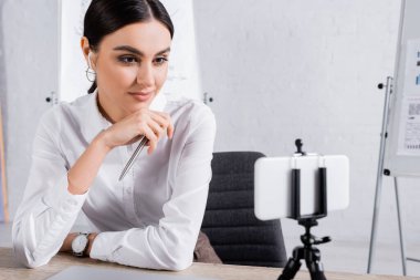 İş kadını kalemle ofisteki selfie çubuğunda bulanık akıllı telefona bakıyor. 