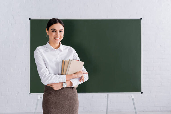 Smiling teacher holding books in classroom 