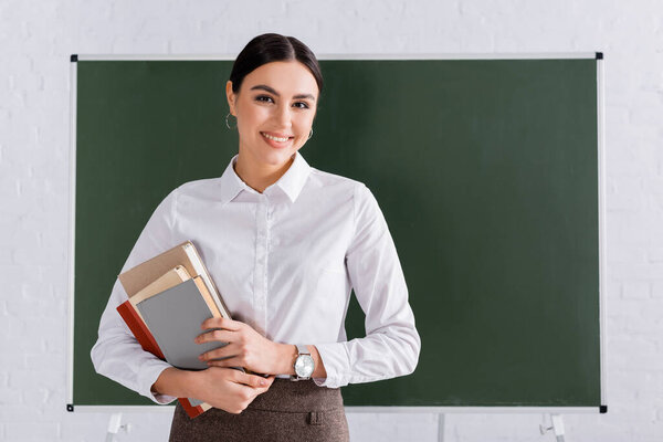 Teacher with books smiling at camera in classroom 