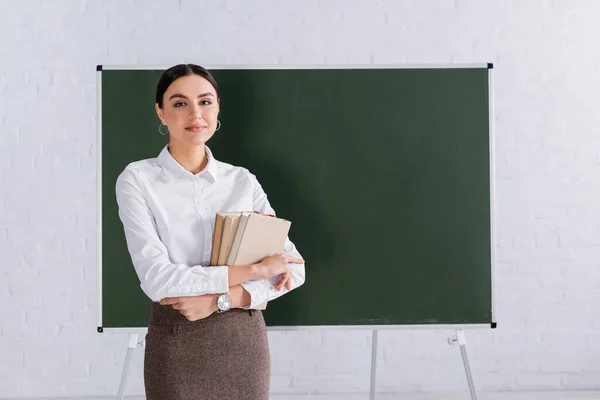Young teacher with books standing near chalkboard in classroom