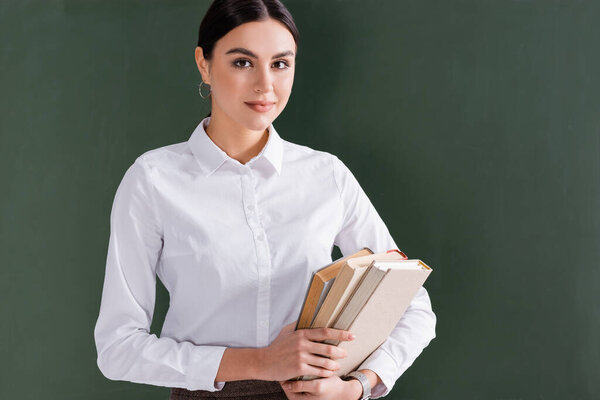 Brunette teacher with books looking at camera near chalkboard 