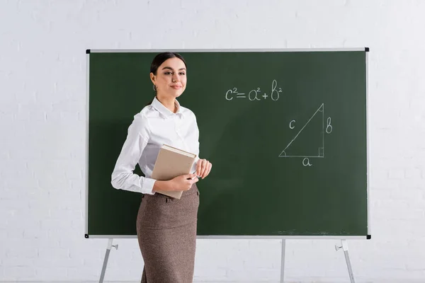 Brunette teacher with book and chalk smiling near equation on chalkboard
