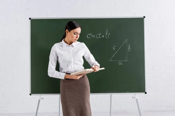 Teacher looking at book near mathematic equation on chalkboard