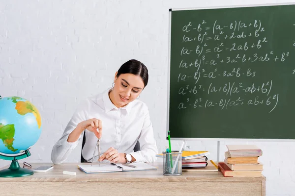 Uśmiechnięty Nauczyciel Trzymający Kompas Rysunkowy Pobliżu Książek Notebooków Stole — Zdjęcie stockowe