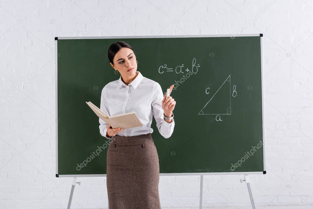 Teacher with book and chalk pointing with finger during lesson in classroom 