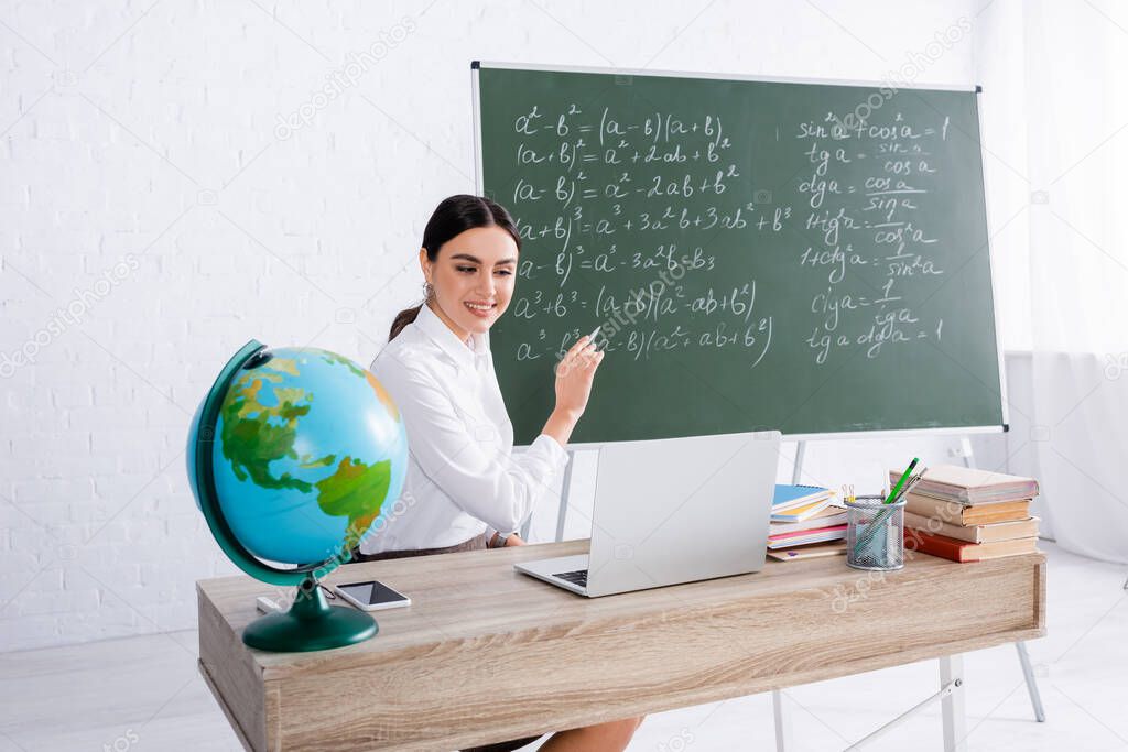 Smiling teacher pointing at chalkboard during online lesson on laptop near globe and books 