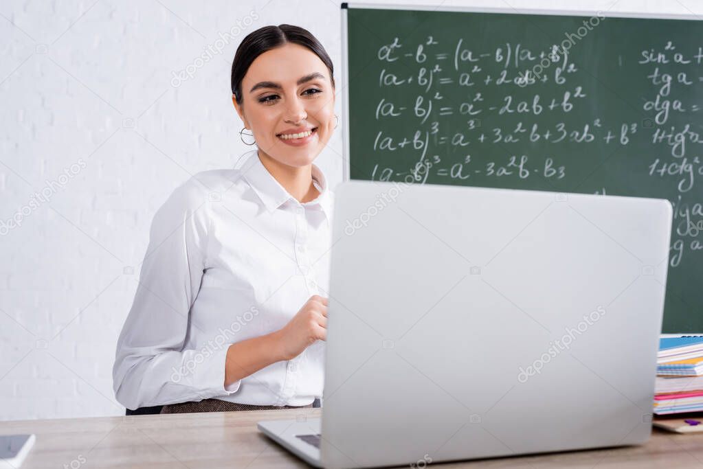 Positive teacher looking at blurred laptop in classroom 